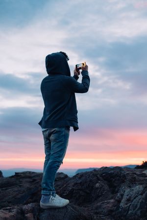 Man on cliff taking photo on phone at sunset