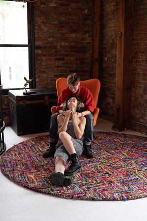 Woman sits on floor and looks up lovingly at her partner sitting in orange chair in urban loft
