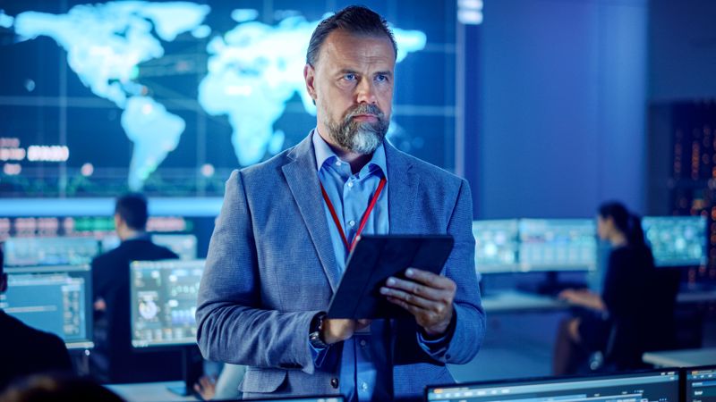 Focused Man in a High-tech Control Room Analyzes Data on a Tablet, Surrounded by Diverse Team Members .