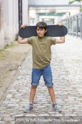 Young boy standing on the street while holding a skateboard behind his head while looking away 4NEyx9