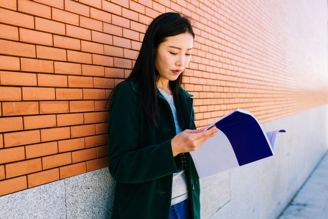 Asian student reading textbook near brick wall