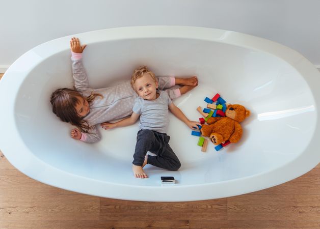Top view children lying in empty bathtub with teddy bear and wooden blocks