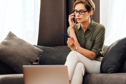 Serious woman on sofa with pillows with cell phone, credit card and laptop