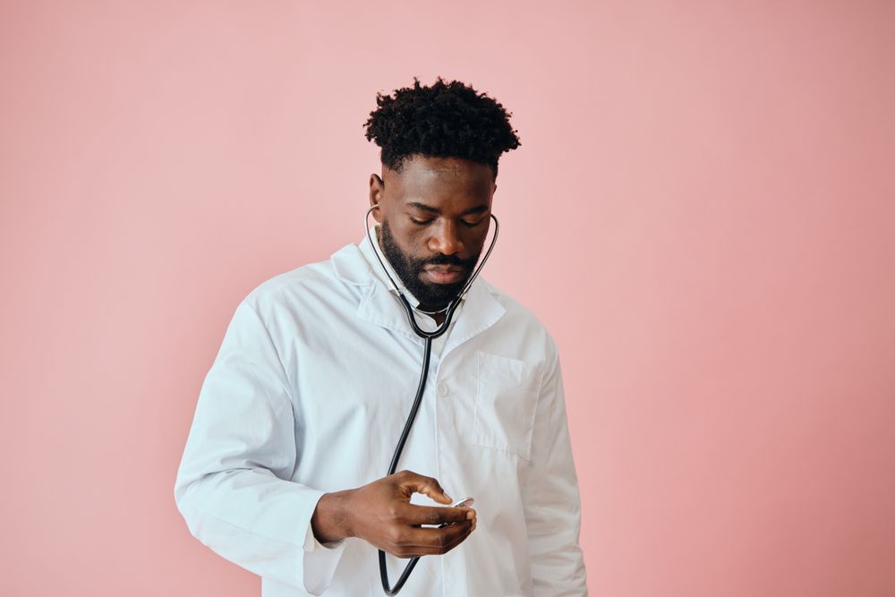Thoughtful Black male doctor in pink studio looking down at his stethoscope