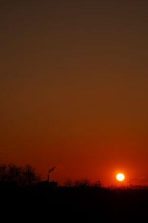 Fiery orange sun sinking behind silhouetted factories releasing smoke