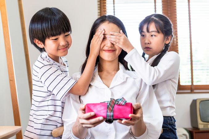 Happy Asian son and daughter covering their mother’s eyes to hide present