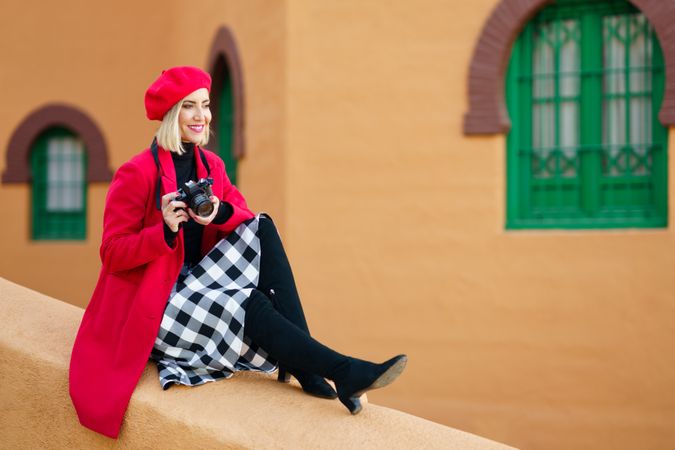 Cheerful female photographer in trendy beret and coat sitting on edge of wall with camera