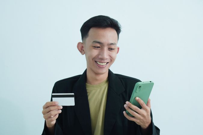Joyful Asian businessman in suit holding blank credit card and smartphone