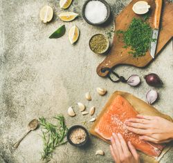 Hand rubbing salt on salmon fillet with fresh herbs, limes, onions, bread board, hand, copy space