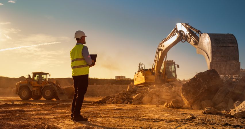 White Male Inspector Using Laptop Computer On Construction Site Of New Real Estate Project. Man Observing Industrial Machinery At Work, Laying Foundation For Apartment Building. Sunny Evening