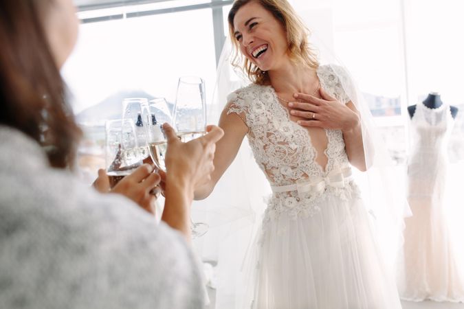 Cheerful young woman in wedding gown toasting champagne with friends in bridal boutique