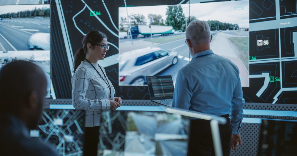 Two Multiethnic Software Engineers Talking Next to a Big Screen in a Modern Monitoring Office with Live CCTV Footage with Traffic Situation.