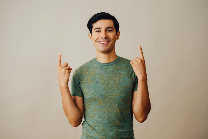 Happy Hispanic male pointing up with both hands in neutral room, copy space