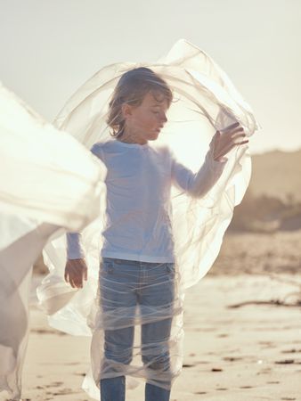 Preteen boy entangled in waving plastic sheet on sandy coast
