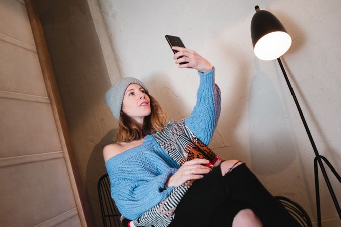 Woman in sweater and hat taking selfie and sitting near wall