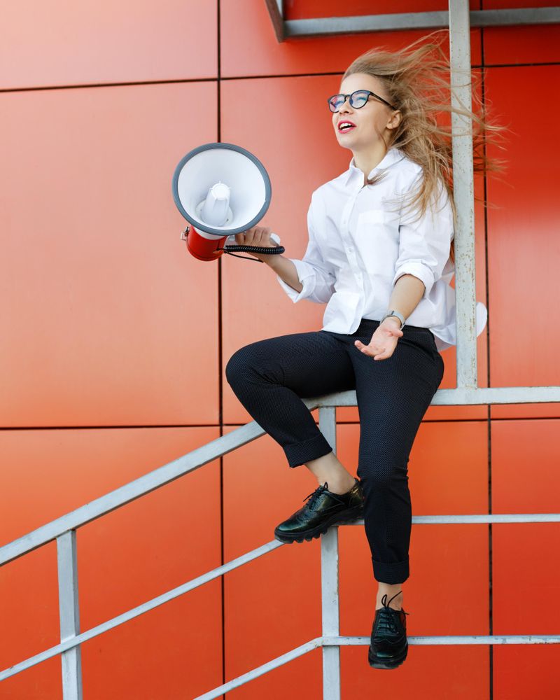 Woman in glasses sitting in front of orange wall with megaphone, vertical