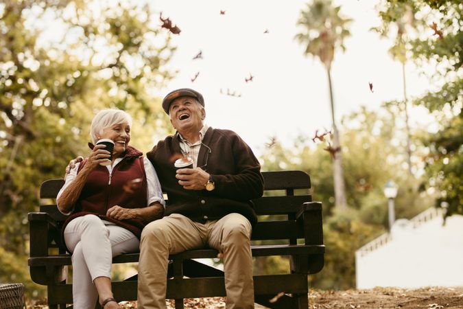 Man and woman with grey hair sitting on park bench with a cup of coffee and laughing