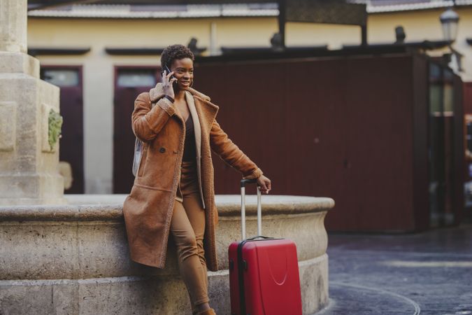 Stylish Black woman talking on smartphone with luggage on street