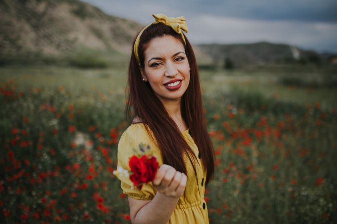 glad adult woman holding rose and looking at camera while standing in meadow