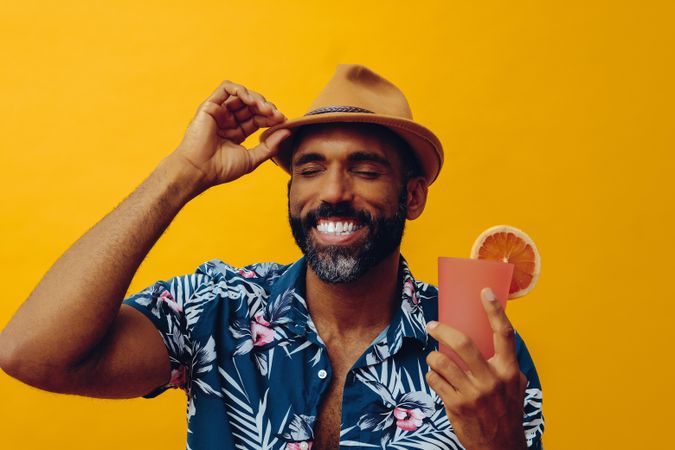 Happy male adjusting hat while wearing colorful shirt and holding cocktail with yellow background