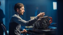 Professional Automotive Engineer in Glasses and Inspection Tools is Testing an Used Electric Engine in a High Tech Laboratory with a Concept Car Chassis.