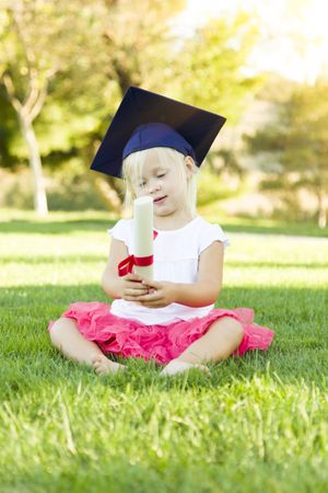 Little Girl In Grass Wearing Graduation Cap Holding Diploma With Ribbon