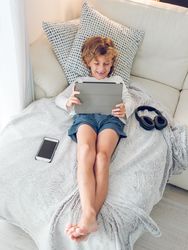 Cheerful boy lying on sofa and watching video on tablet