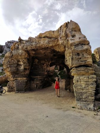 Woman in red dress standing beside arched rock formation in Sicily, Italy