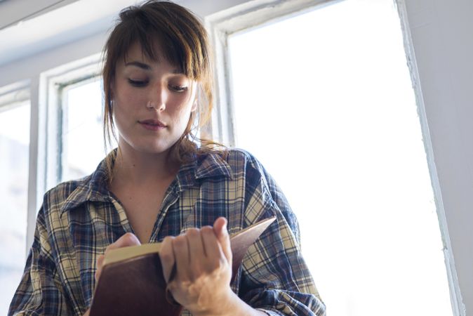 Female in flannel shirt writing in notebook in bright room