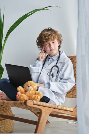 Thoughtful boy in medical uniform with laptop and toy bear