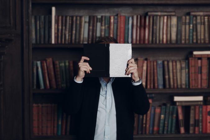 Man in dark suit holding a book to his face standing against bookshelves