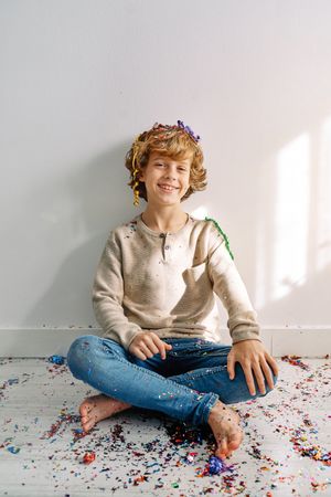 Positive cute boy sitting on floor covered with confetti