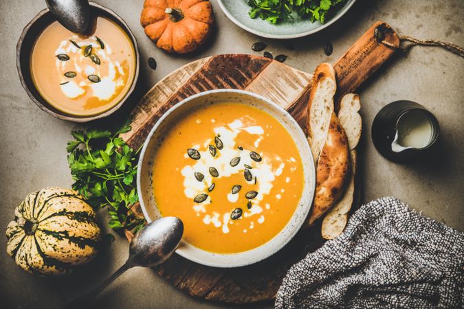 Two bowls of pumpkin soup with garnishes, bread, cream, squash, on concrete surface