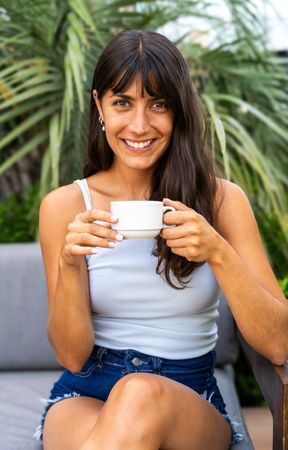 A woman is lookintg at camera while sitting on a couch and holding a white cup. She is smiling and she is enjoying her coffee
