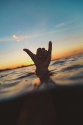 Hand with shaka gesture between water surface and sky at sunset