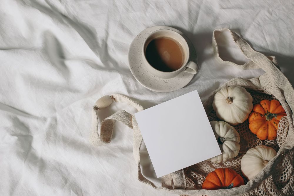 Autumn stationery mockup of blank square greeting card, pumpkins in mesh bag on linen table cloth background and cup of coffee