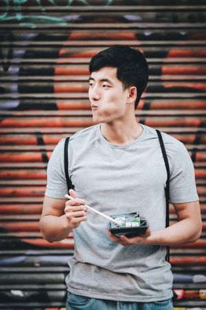 Asian man eating takeaway food with chopsticks on city street