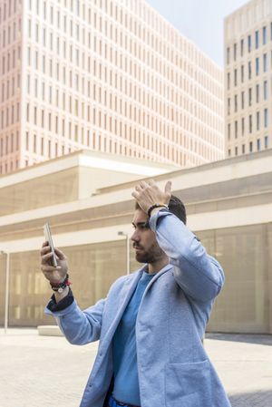 Man in blazer standing outside checking phone with hand on forehead