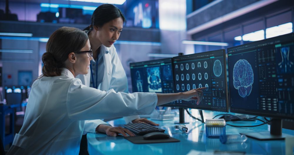 Medical Hospital Research Laboratory: White And Asian Female Scientists Talking And Using Desktop Computer With MRI Scans Of Human Brain.
