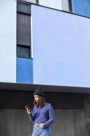 Female in wool hat and sweater checking phone outside blue building, vertical