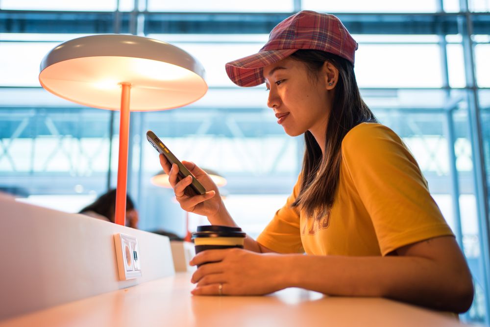 Asian woman browsing smartphone while drinking coffee at table under lighting lamp