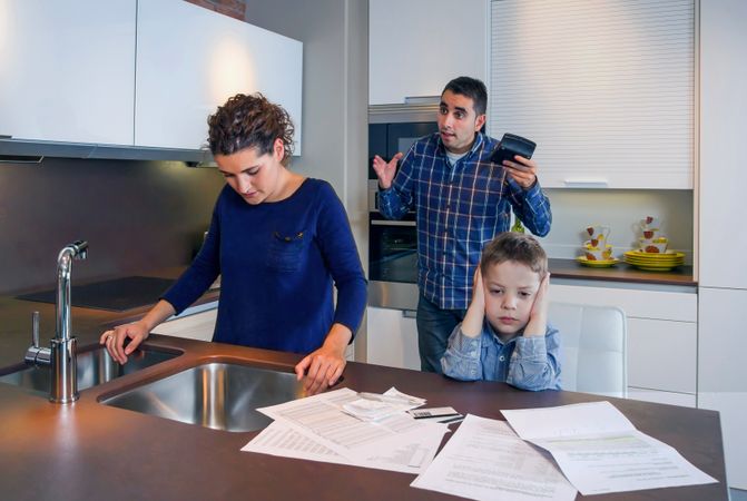 Couple arguing in front of sad son with bills strewn on the kitchen counter