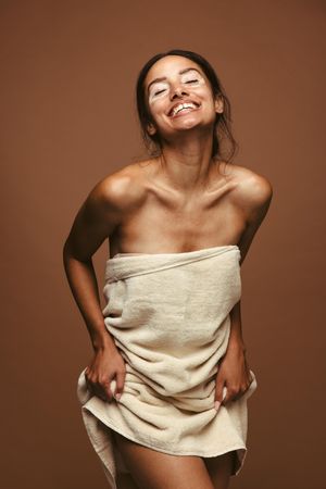 Cheerful woman standing against brown background wearing bath towel
