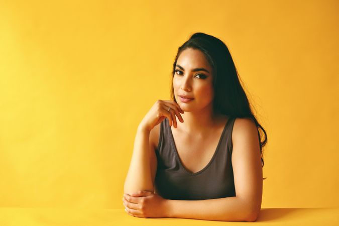 Pensive Hispanic woman looking at camera resting chin on her hand and sitting in yellow room
