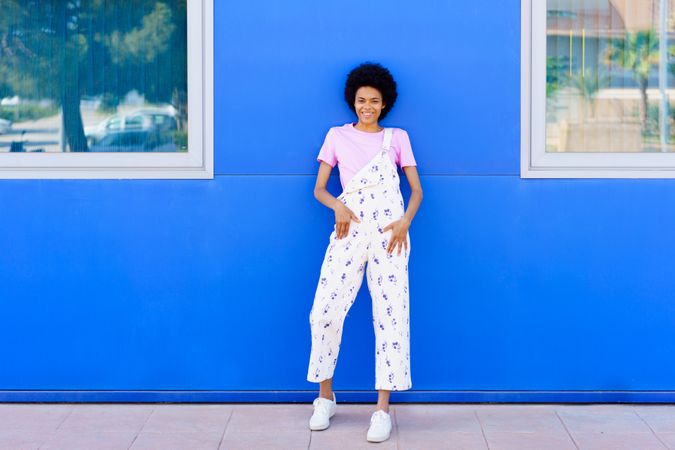 Happy woman in floral coveralls leaning against outside of blue building