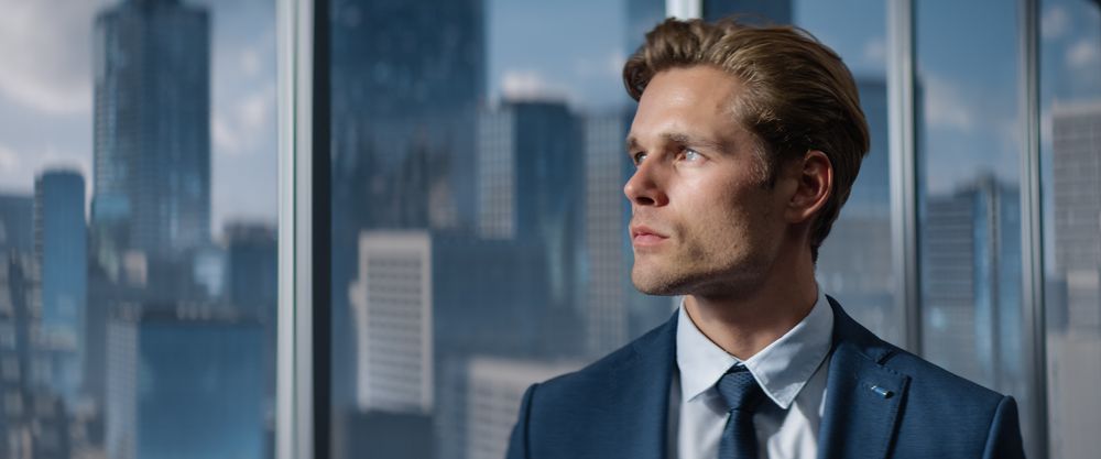 Portrait of Successful Businessman in a Tailored Suit Standing in Office Looking out of the Window on Big City with Skyscrapers.