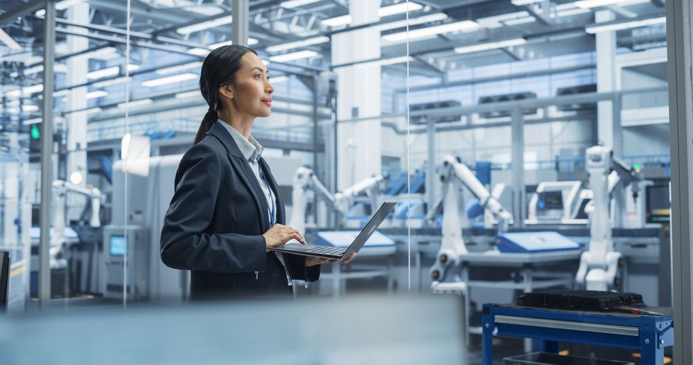 Asian Female Engineer Standing Next to a Window in Office, Using Laptop Computer at an Electronics Factory. Machines are Undergoing Maintenance, Specialist Using an Online Software