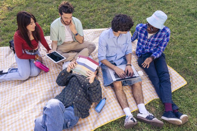 Group of young people sitting on blanket with books laptop tablet outdoor