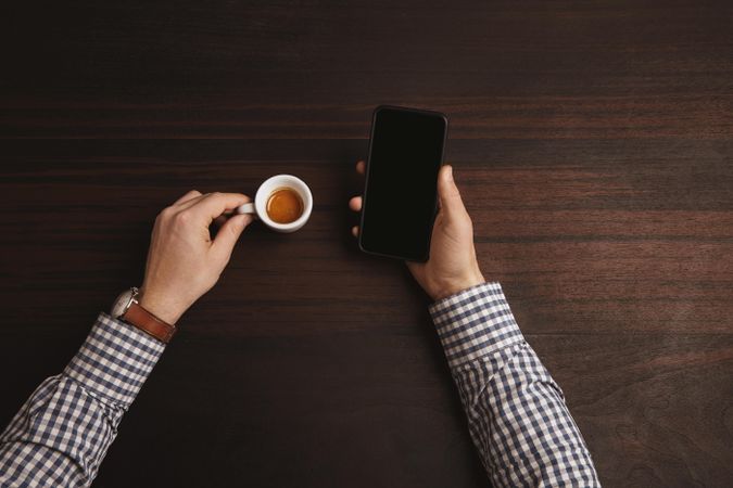 Man wearing plaid shirt and leather watch holding phone in one hand, espresso shot in the other