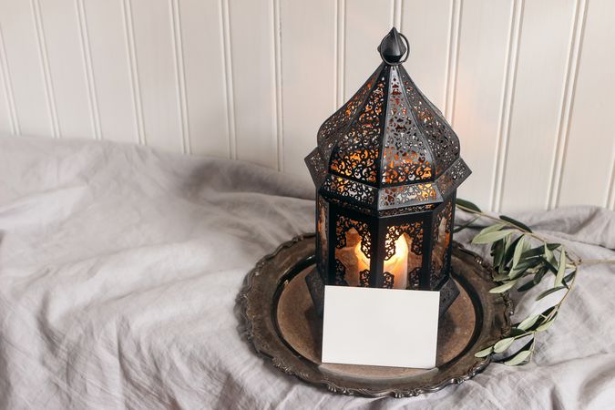 Ramadan Kareem holiday still life. Dark Moroccan lantern with olive tree branches on silver tray. Linen table cloth. Blank greeting card, invitation mockup on wooden table. Festive photo. Top view